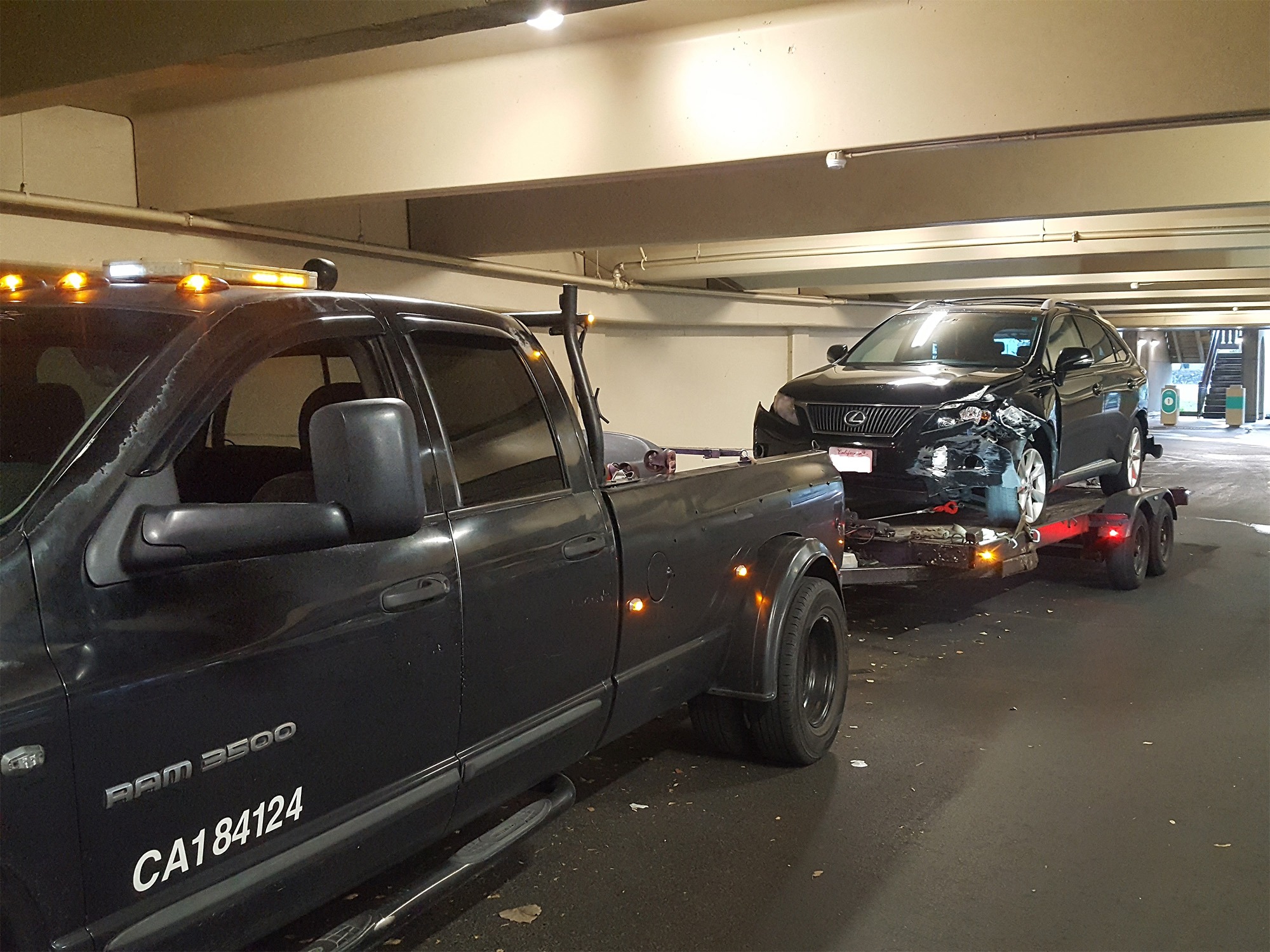 A black tow truck pulls a damaged black SUV up a trailer in a parking garage, indicating a vehicle recovery or towing operation.
