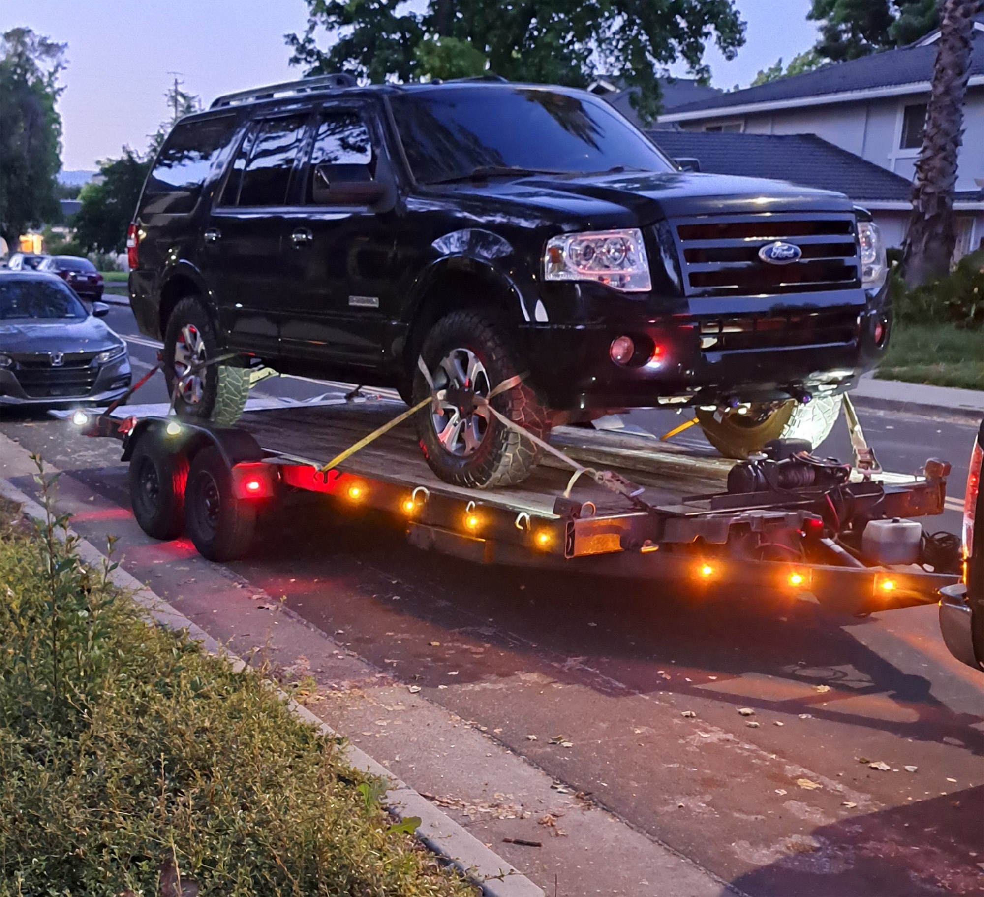 A black Ford SUV is secured on a trailer, illuminated by lights, parked on a suburban street during twilight.