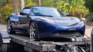 A blue Tesla Roadster is parked on a flatbed trailer surrounded by palm trees, showcasing its sleek design and sports car features.