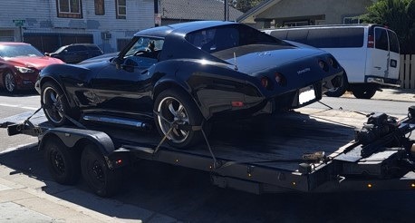 A black classic Corvette sports car is loaded onto a trailer on a sunny street, indicating transport or restoration purpose.