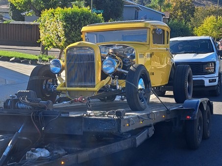 Classic yellow hot rod on a trailer, showcasing its vintage design and exposed engine, surrounded by a suburban neighborhood.
