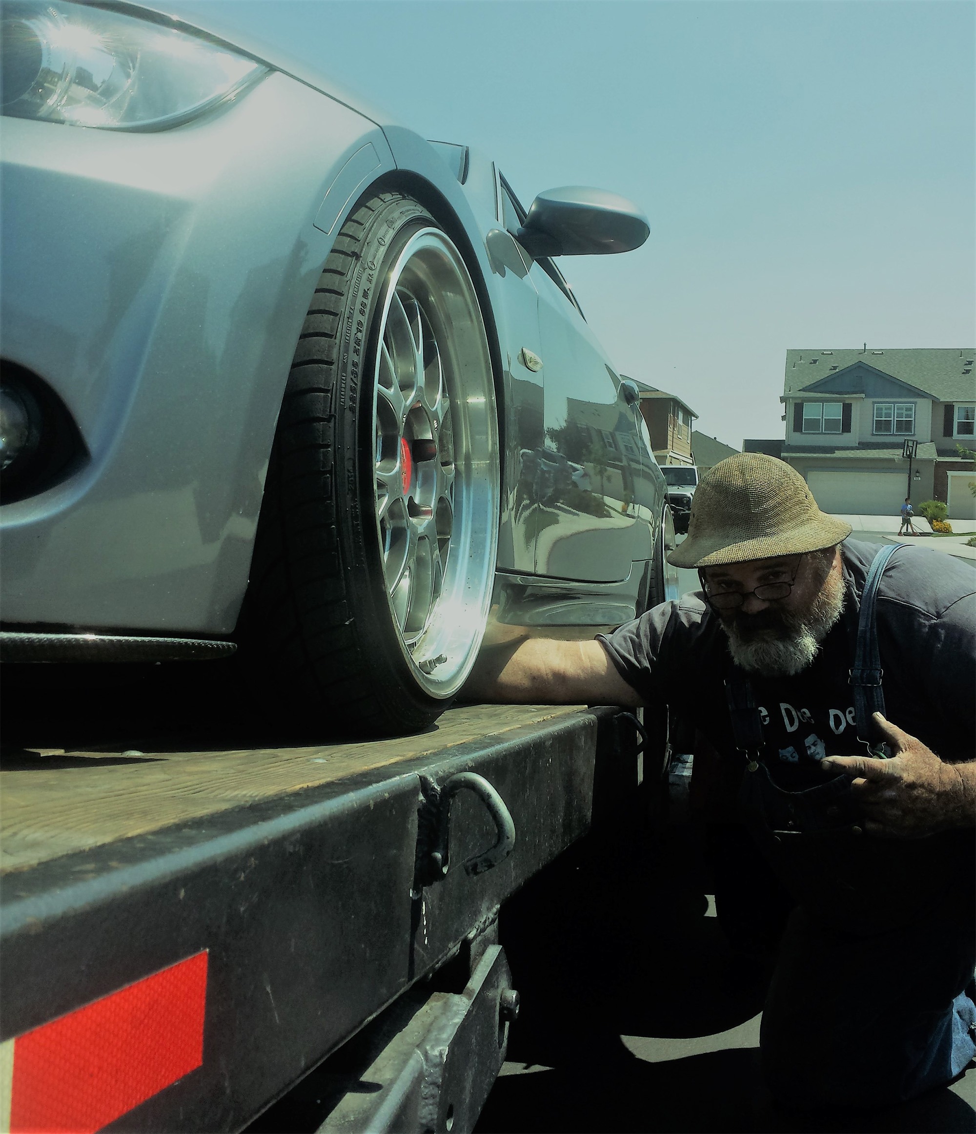 A mechanic inspects a lowered silver car on a tow truck ramp, focusing on the wheel and tire. The background features suburban homes, indicating a roadside assistance context.
