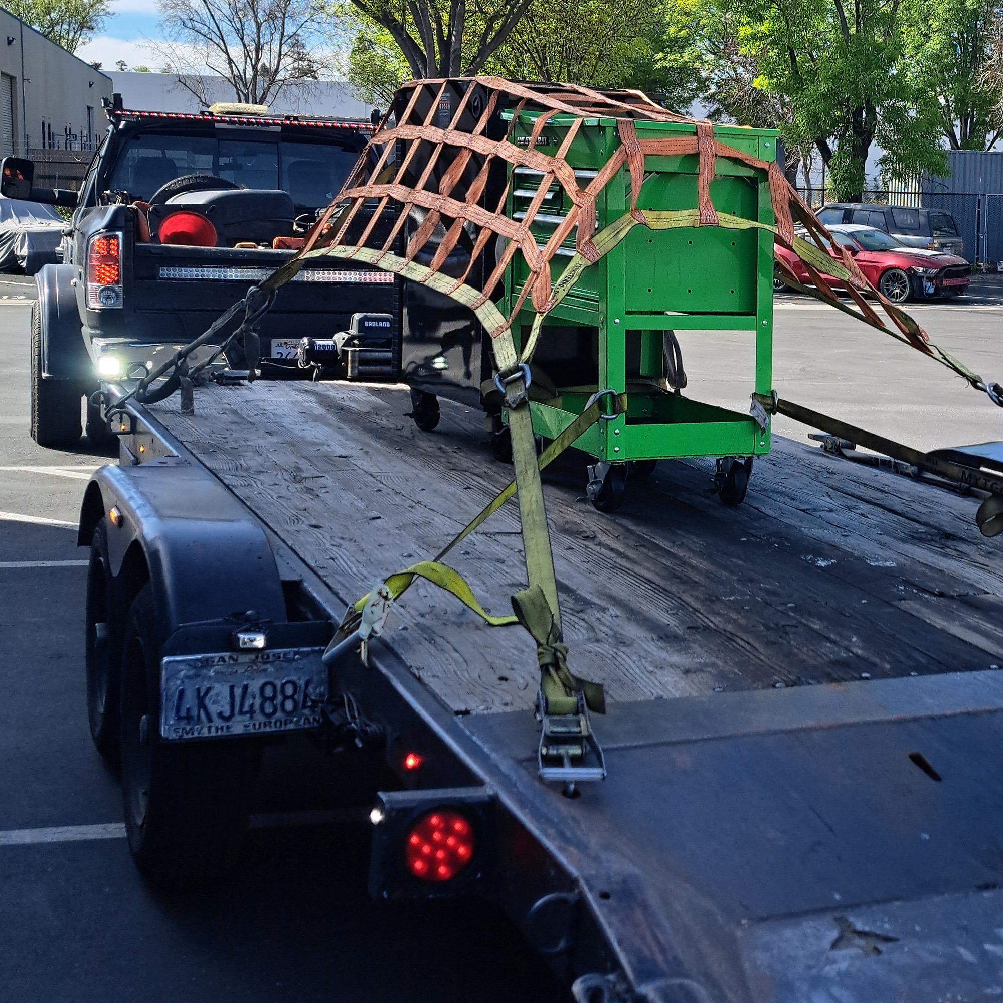 A green tool cart secured with straps on a flatbed trailer, attached to a pickup truck. The scene illustrates equipment transportation.