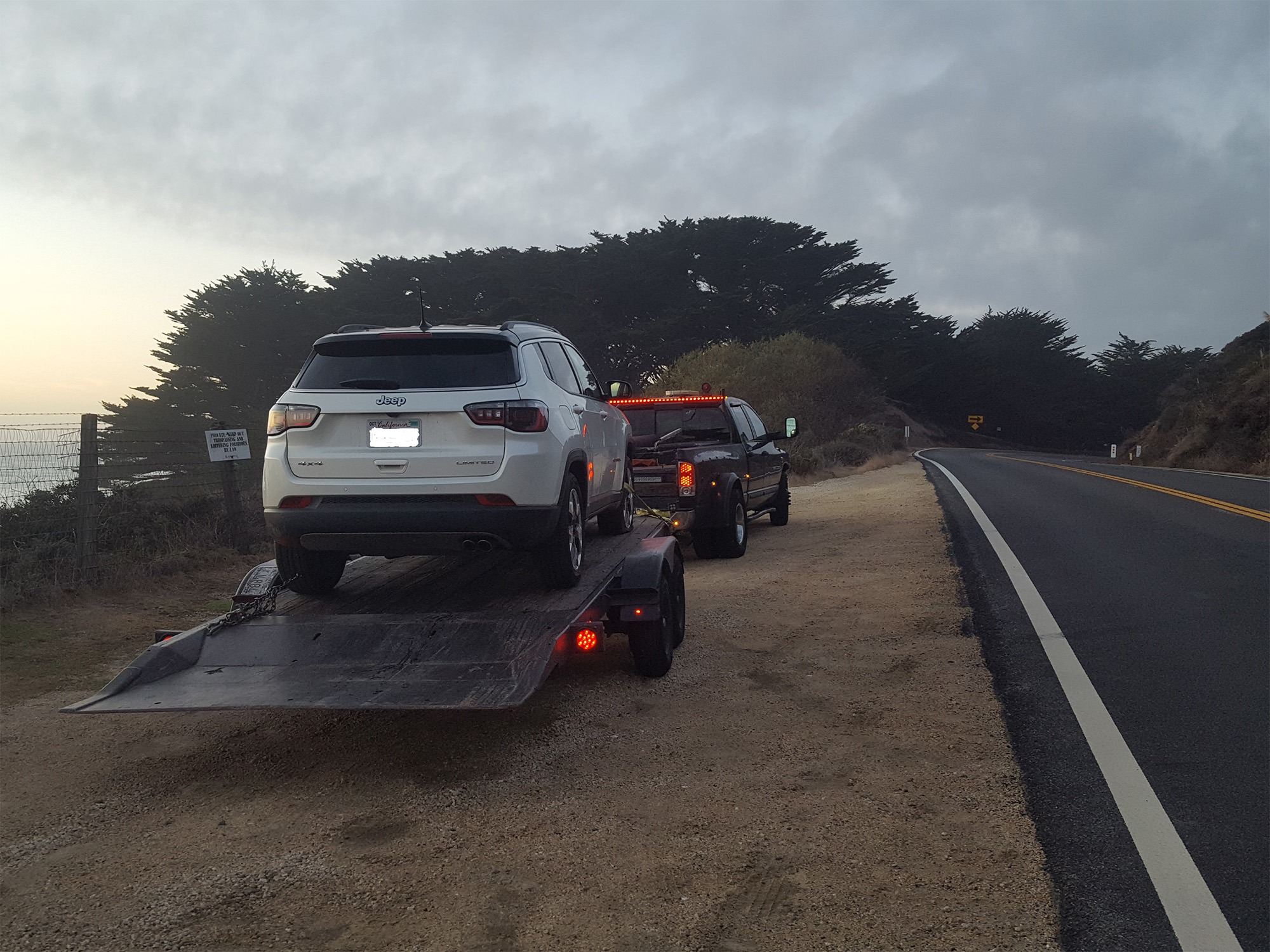 A white Jeep is loaded onto a flatbed trailer beside a coastal road, with a pickup truck nearby. The setting is early morning with a cloudy sky.