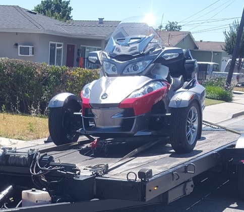 A silver and red Can-Am Spyder motorcycle on a tow truck, positioned on a suburban street with houses in the background.