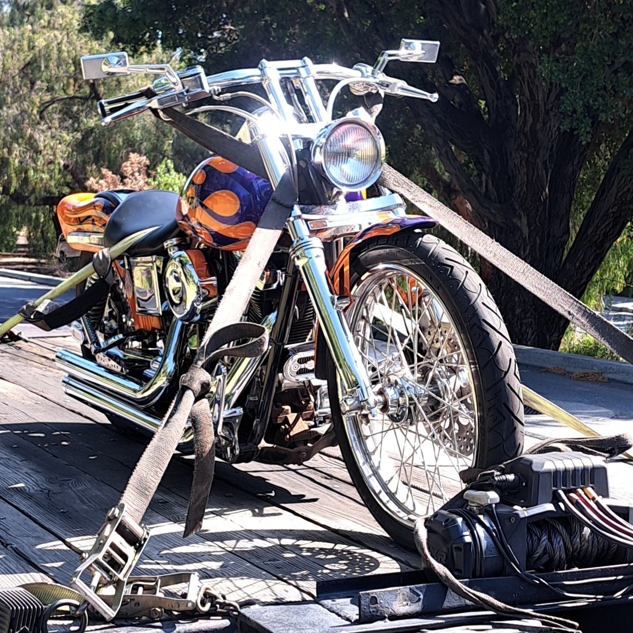 Custom motorcycle secured on a trailer with straps, featuring a vibrant orange and purple paint job. Surrounded by greenery, it highlights its unique design.